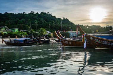 Koh Phi Phi, Tayland - Kasım 2018: şafak vakti, sabah güneşi gökyüzünde, yemyeşil ağaçlar ve palmiye ağaçları arka planda adanın kenarında dizilmiş tekneler plaj denize yakın evler
