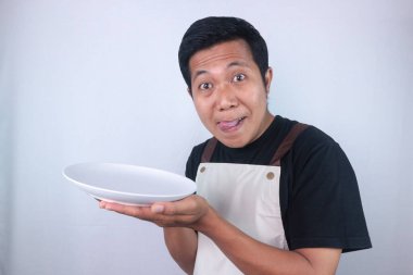 Funny expression asian young man shows empty white plate while stuck out his tongue. Showing delicious dish. Advertisement concept