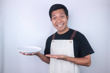 Smiling asian man chef or waiter shows empty white plate, presenting something. Isolated white background