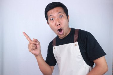 Funny shocked face expression young asian man wearing apron pointing right side, empty space. advertisement concept
