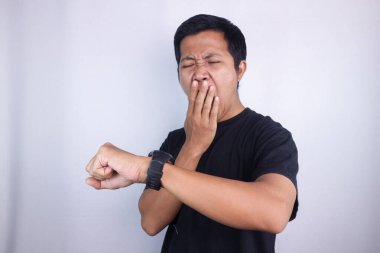 Tired Asian man yawning covering mouth looks at his watch, bed time, rest. isolated on white background