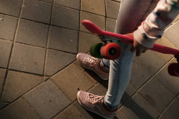 Teenager girl holding penny skateboard. Close up of Pink skateboard and female legs in sneakers on street on sunrise.