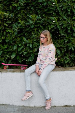  pembe kap ve kaykay parkı ile sarışın kız portresi. Gözlük açık havada sokakta oturan genç kız. Çocukluk, yaz, tatil, spor 