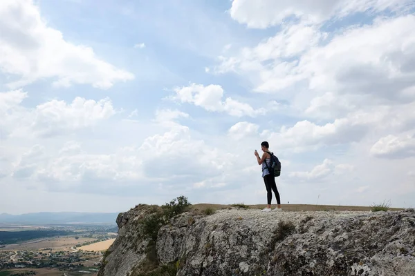 年轻女子徒步旅行者站在悬崖边上使用手机 在山顶拍照的女游客 远足概念 — 图库照片