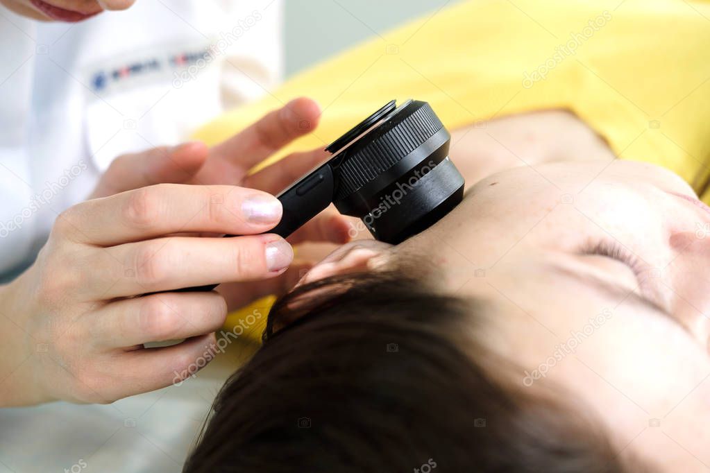 Female dermatologist using a professional dermatoscope while doing skin examination, Checking benign moles on face. Dermatologist examining birthmarks and moles on a female patient. Dermatology Clinic