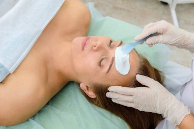 Yüz soyma maskesi, spa güzellik tedavisi, cilt bakımı. Spa salonunda güzellik uzmanı tarafından yüz bakımı yaptıran kadın. bir kız hastanın yüzüne krem maskesi uygulayan ometolog. Kozmetoloji. Yakın çekim