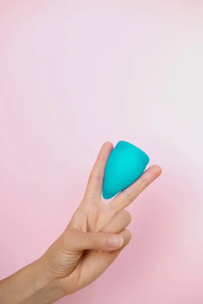 Woman holding on two finger blue menstrual cup on pink background. Feminine hygiene alternative product instead of tampon during period. Menstruation, critical days, women periods. Zero waste