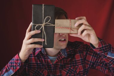 Ekose gömlekli adam yüzünü kapatıyor. Kırmızı arka planda Noel hediyesi. Kış tatilleri için hediyeli erkek eller. Noel, yeni yıl, alışveriş, tatil hazırlıkları. Kapat.
