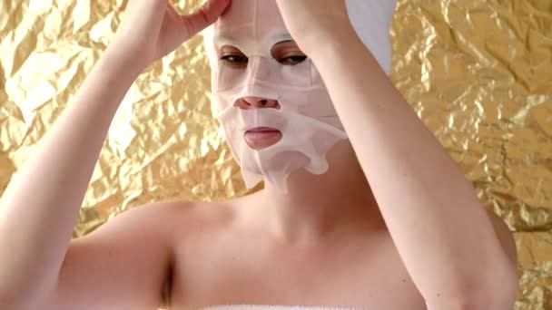 年轻女人在金色的背景上涂上化妆品织物面膜 面部保湿面膜用木炭 温泉美容治疗 靠近点 — 图库视频影像