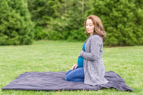 Glücklich schwangere Kaukasierin entspannt sich in einem Park und hält die Hände auf ihrem Bauch. — Stockfoto