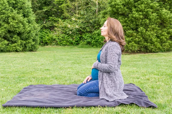 Glücklich schwangere Kaukasierin entspannt sich in einem Park und hält die Hände auf ihrem Bauch. — Stockfoto