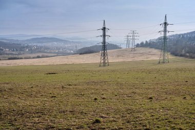 Manzaralı manzara parkındaki metal çerçeve aktarım kuleleri üzerindeki iletken kablolar vasıtasıyla dağlık bölgedeki küçük bir kasabaya elektrik tedarik ediliyor. Çayır dağlarında mavi gökyüzünün altındaki eski yüksek voltajlı elektrik şebekesi direkleri. Litomerice, Çek Cumhuriyeti, Avrupa.
