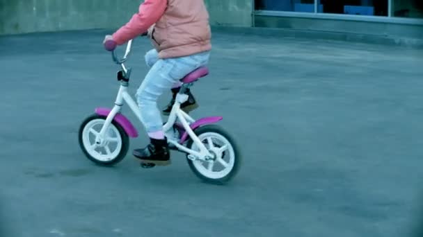 Una madre sonriente y riente empuja a su hija hacia adelante en un cálido día de primavera, cuando le enseña a andar en bicicleta por la acera de la ciudad cerca de un parque verde. . — Vídeo de stock