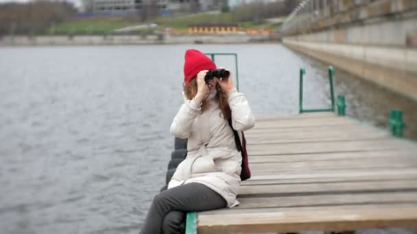 Una hermosa mujer con una chaqueta y un sombrero rojo está de pie en el muelle y mirando a través de binocular — Vídeo de stock