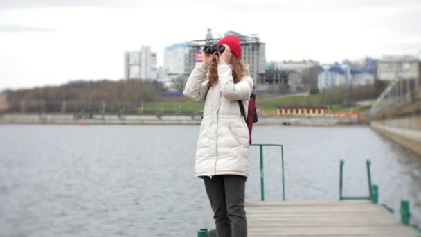 En vacker kvinna i en jacka och en röd hatt står på bryggan och tittar genom kikaren — Stockvideo