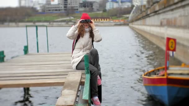 En vacker kvinna i en jacka och en röd hatt står på bryggan och tittar genom kikaren — Stockvideo