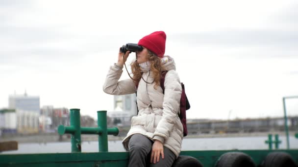 Uma mulher bonita em uma jaqueta e um chapéu vermelho está de pé na doca e olhando através binocular — Vídeo de Stock