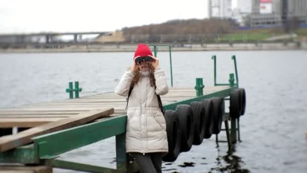 En vacker kvinna i en jacka och en röd hatt står på bryggan och tittar genom kikaren — Stockvideo