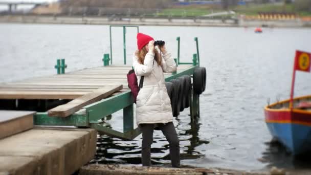 Una hermosa mujer con una chaqueta y un sombrero rojo está de pie en el muelle y mirando a través de binocular — Vídeos de Stock