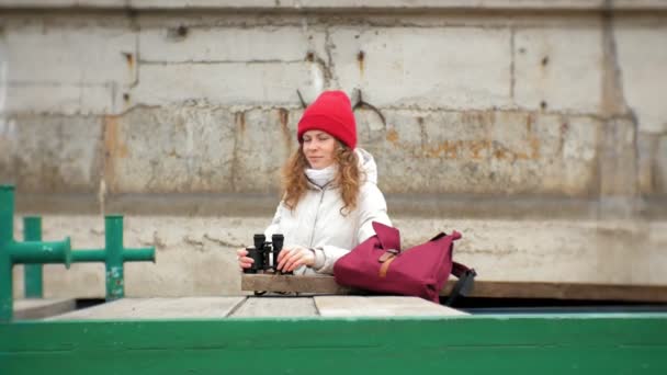 Una hermosa mujer con una chaqueta y un sombrero rojo está de pie en el muelle y mirando a través de binocular — Vídeos de Stock