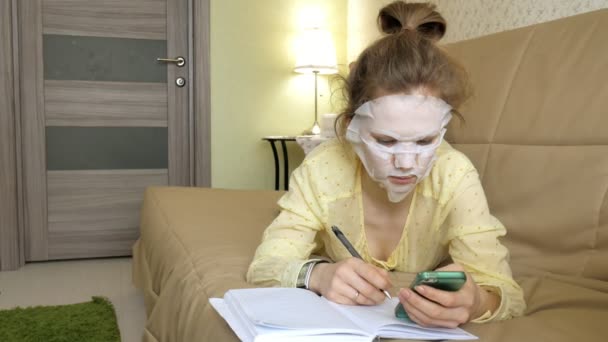 Mujer joven haciendo máscara facial con máscara de limpieza, clics en el sofá con teléfono inteligente en casa — Vídeos de Stock