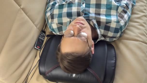 Hombre se encuentra en el sofá en casa, bajo la cabeza de un masajeador, estado relajado — Vídeo de stock