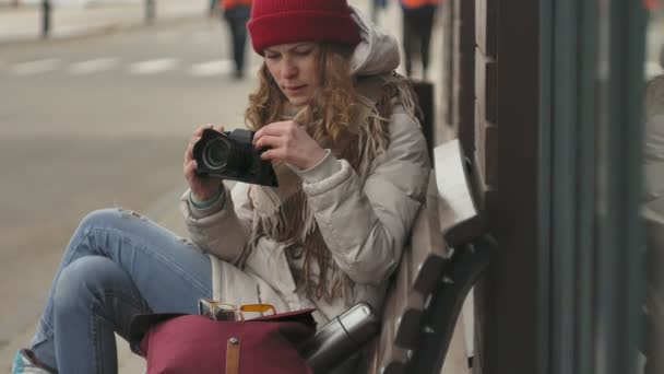 Joven hermosa mujer en un sombrero rojo con ropa de abrigo deportivo y rodillos, sentado en un banco de madera y tomando fotos en una cámara vintage — Vídeos de Stock