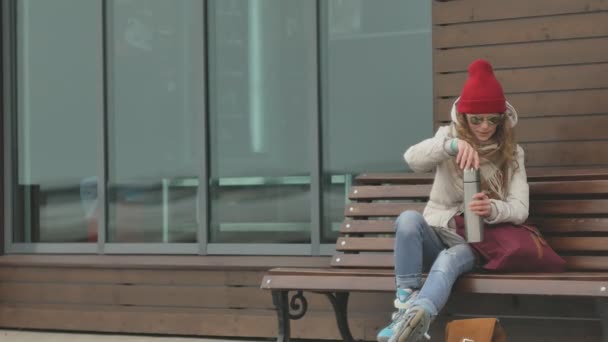 Mladá krásná žena v červeném klobouku na sobě teplé sportovní oblečení a válečky, seděl na dřevěné lavičce pití čaje z termosky — Stock video