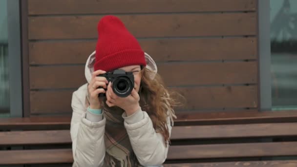 Jovem mulher bonita em um chapéu vermelho vestindo roupas e rolos quentes esportivos, sentado em um banco de madeira e tirando fotos em uma câmera vintage — Vídeo de Stock