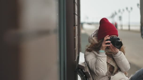 Jeune belle femme au chapeau rouge portant des vêtements chauds et sportifs et des rouleaux, assise sur un banc en bois et prenant des photos sur un appareil photo vintage — Video