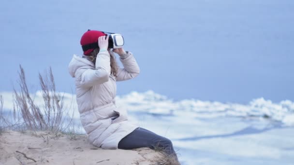 Kırmızı bir şapka çekici genç kadın turist oturur bir sahilde ve sanal gerçeklik kask sahiptir — Stok video