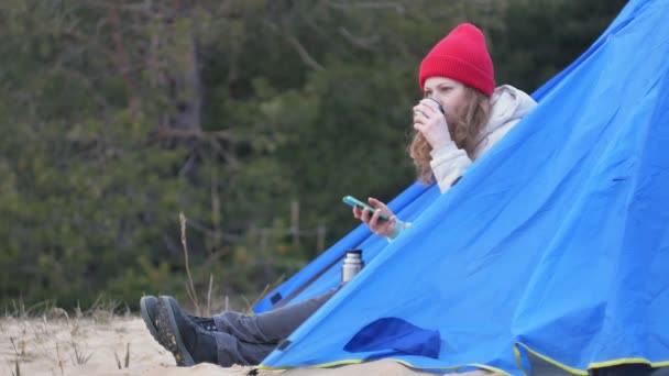 Attraktive junge Touristin mit rotem Hut sitzt in einem Touristenzelt und trinkt Tee aus einer Thermoskanne — Stockvideo