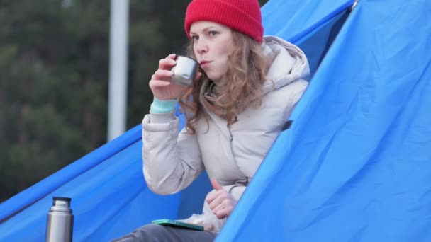 Jovem turista atraente em um chapéu vermelho senta-se em uma tenda turística e bebe chá de uma garrafa térmica — Vídeo de Stock