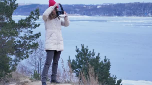 Attractive young woman tourist in a red hat sits in a on the shore and enjoys a virtual reality helmet — Stock Video