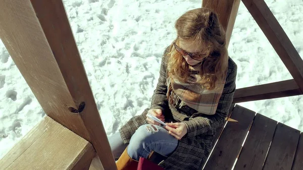 Una joven se sienta en una veranda de madera, tocando silenciosamente el avión de papel de origami con los dedos y poniéndolo de rodillas. Chica sentada al aire libre con aviones de aerogamia blanca, vista cercana , — Foto de Stock