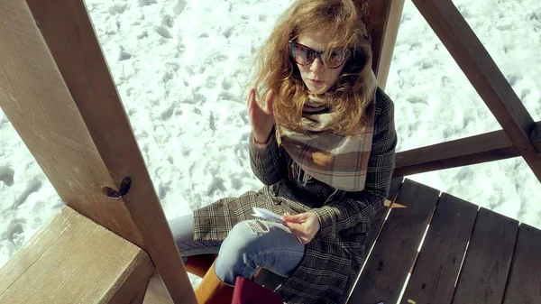Una giovane donna si siede su una veranda di legno, toccando silenziosamente l'aereo di carta origami con le dita e mettendolo in ginocchio. Ragazza seduta all'aperto con aereo aerogamia bianco, vista da vicino , — Foto Stock