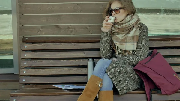 Giovane donna che beve tè da una bottiglia di thermos in un parco primaverile seduta su una panchina — Foto Stock