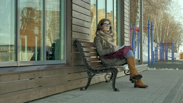 Молодая, красивая, милая женщина, сидящая на скамейке в парке, в руках своего блокнота. На ней пальто. Она делает заметки и выглядит очень романтично . — стоковое фото