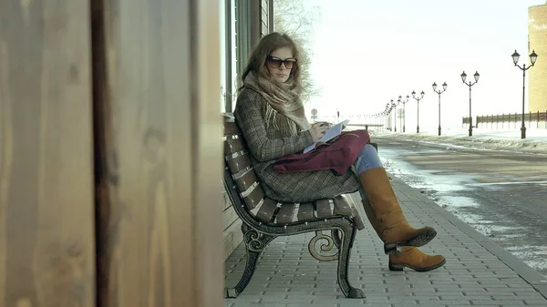 Junge, schöne, schöne Frau, die auf einer Bank im Park sitzt und ihr Notizbuch in der Hand hält. Sie trägt einen Mantel. sie macht Notizen und sieht sehr romantisch aus. — Stockfoto