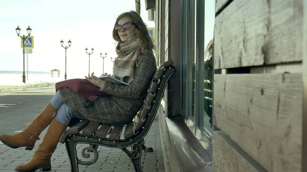 Joven, hermosa, encantadora mujer sentada en el banco en el parque, en las manos de su cuaderno. Lleva un abrigo. Ella toma notas y se ve muy romántica . —  Fotos de Stock