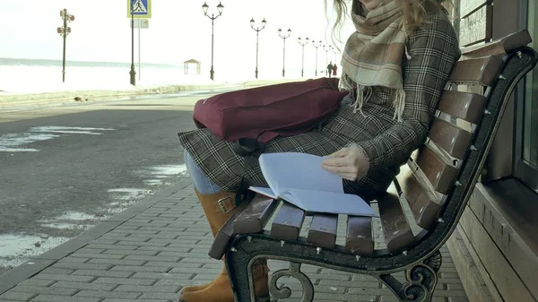 Junge, schöne, schöne Frau, die auf einer Bank im Park sitzt und ihr Notizbuch in der Hand hält. Sie trägt einen Mantel. sie macht Notizen und sieht sehr romantisch aus. — Stockfoto