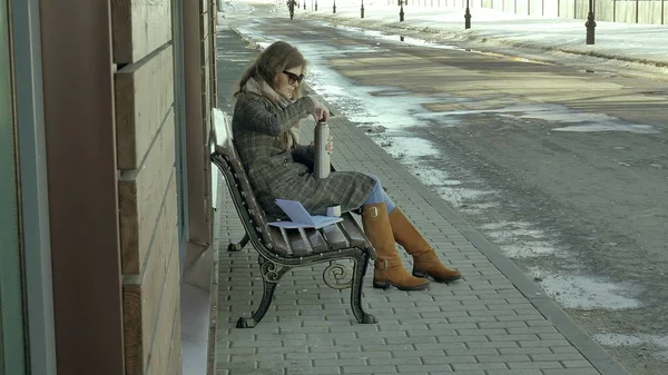 Jonge vrouw drinken thee uit een thermos fles in een park van de lente zittend op een bankje — Stockfoto