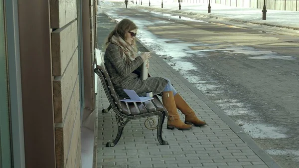 Mujer joven bebiendo té de una botella de termo en un parque de primavera sentado en un banco — Foto de Stock