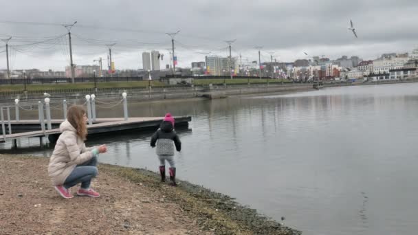 Boldog és gyönyörű anya és lánya etetés sirályok a strandon, a folyó partján — Stock videók