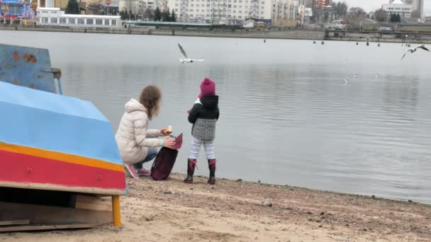 Szczęśliwe i piękne matka i córka są karmienia mewy na plaży, nad brzegiem rzeki — Wideo stockowe