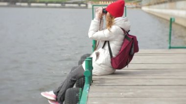 Güzel bir kadının bir ceket ve kırmızı bir şapka iskelede duran ve dürbün ile seyir