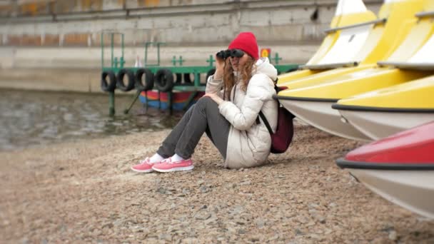 Una hermosa mujer con una chaqueta y un sombrero rojo está de pie en el muelle y mirando a través de binocular — Vídeos de Stock