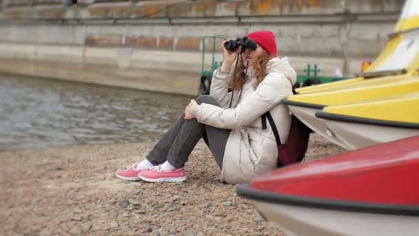 Una hermosa mujer con una chaqueta y un sombrero rojo está de pie en el muelle y mirando a través de binocular — Vídeos de Stock