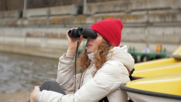 A beautiful woman in a jacket and a red hat is standing on the dock and looking through binocular — Stock Video