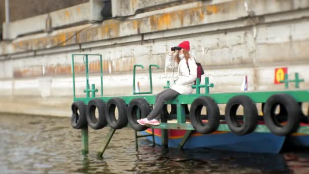 Una bella donna in giacca e cappello rosso è in piedi sul molo e guardando attraverso binocolo — Video Stock